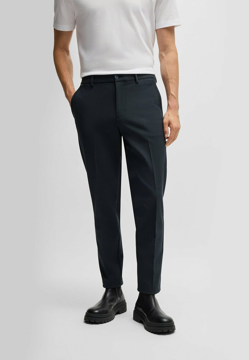 Dark tailored trousers with a slim fit, featuring a smooth fabric, straight legs, and a single button closure. Paired with black boots.