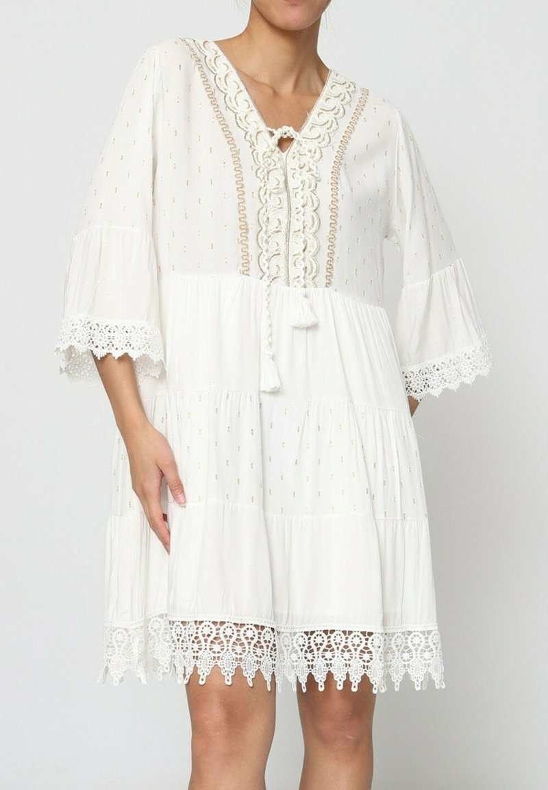 Robe blanche avec un col en V, un corsage brodé et des bordures en dentelle. Comprend des manches trois-quarts et un design en étages avec un détail à pompons.