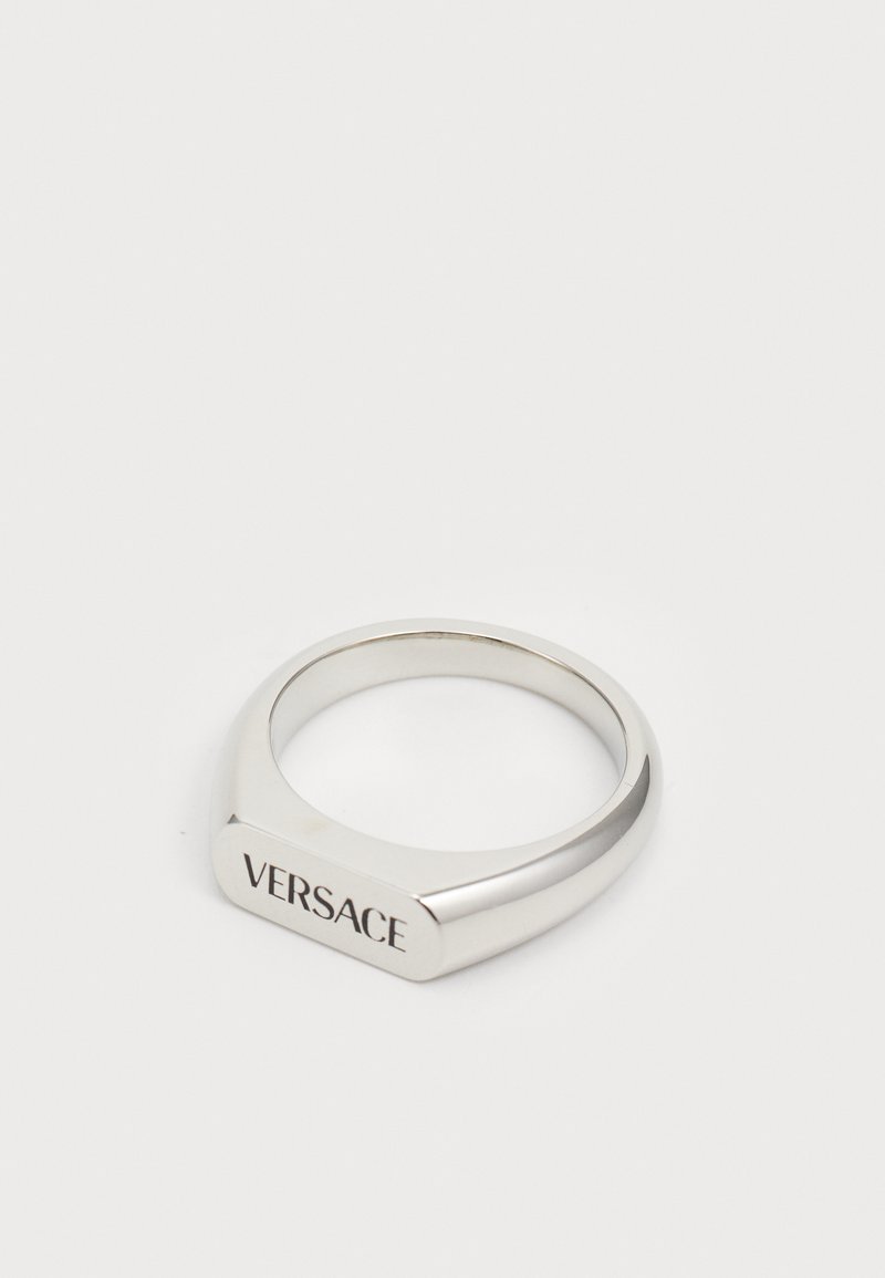 Zilveren ring met een geometrische vorm, voorzien van een plat bovenstuk gegraveerd met "VERSACE." Gladde textuur en gepolijste afwerking.