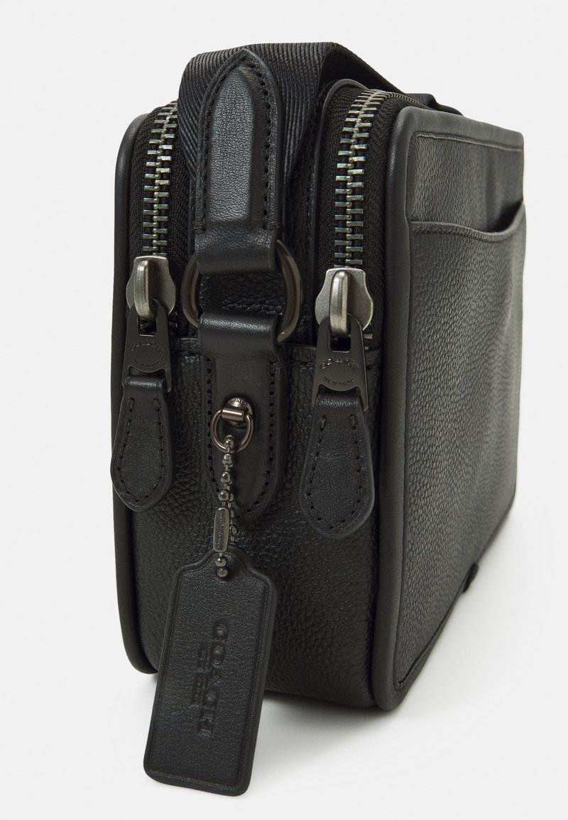 Sac en cuir noir avec une finition texturée, double zip en argent et une petite étiquette suspendue. Forme rectangulaire compacte avec une poche latérale.