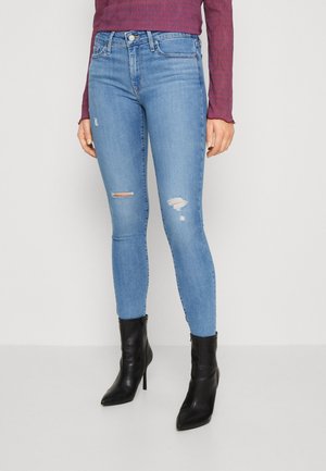Femme portant un jean skinny bleu clair déchiré, des bottines noires à talons hauts et un haut à manches longues bordeaux, debout devant un fond uni.