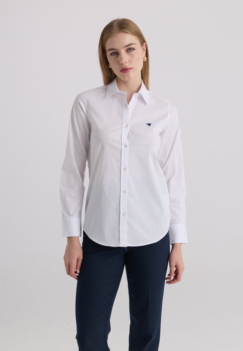 Jeune femme aux cheveux blonds raides portant une chemise blanche boutonnée et un pantalon foncé, debout devant un fond clair uni.