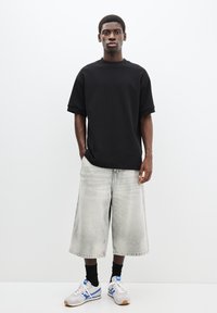 Cămașă neagră cu mânecă scurtă, croială lejeră, asortată cu pantaloni scurți din denim gri deschis. Modelul poartă adidași albi cu accente albastre și șosete negre.