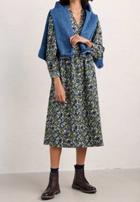 Šaty s květinovým vzorem v tmavě zelené barvě, s dlouhými rukávy a ke kolenům dlouhým lemem. Džínová bunda volně přehozená přes ramena. Hnědé boty na šněrování.