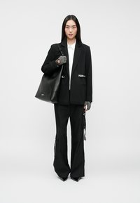 Γυναίκα που φοράει μαύρο σακάκι και παντελόνι, λευκό πουκάμισο, γκρι γάντια χωρίς δάχτυλα, κρατώντας μια μεγάλη μαύρη τσάντα, standing on a white background.