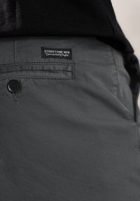 Shorts en tissu gris au toucher lisse, dotés d'une fermeture à bouton avant et d'une petite étiquette noire portant l'inscription "STREETONE MEN" sur la ceinture.
