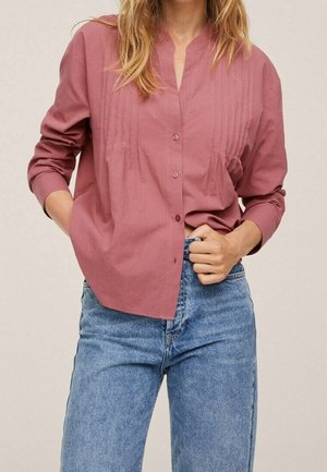 Kvinde iført en støvet rosa langærmet skjorte med knapper og lodrette plisseringer, stoppet ned i højtaljede blå jeans, med den ene hånd i lommen.