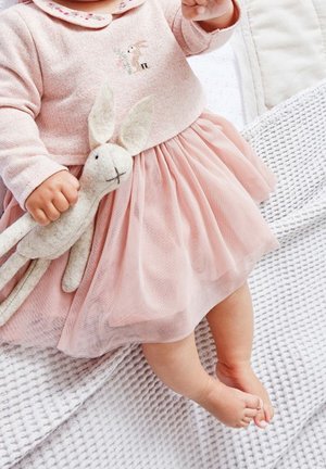 Bébé en robe rose tenant un jouet lapin gris doux, assis sur une couverture blanche texturée avec les pieds nus visibles.
