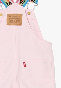 Pettorina di denim rosa con spalline regolabili arcobaleno, dotata di bottoni dorati, etichetta in pelle e due tasche anteriori. Etichetta rossa con logo visibile.