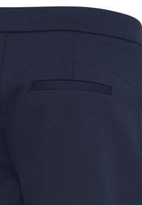 Gros plan d’un pantalon bleu marine montrant une poche passepoilée arrière avec une ceinture en tissu lisse et des détails de couture.