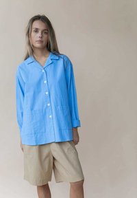 Lichtblauw overhemd met lange mouwen en kraag, twee voorzakken en witte knopen, gecombineerd met beige shorts van een zachte stof.