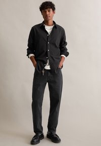 Črna srajca z gumbi, pod njo bela majica s kratkimi rokavi, temno sivi hlače in črne čevlje, stoji ob nevtralni ozadju.