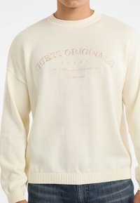 Cremefarbener gestrickter Pullover mit gerippten Bündchen und Saum. Verfügt über den gestickten Text "GUESS ORIGINALS" und dekorative Sterne.
