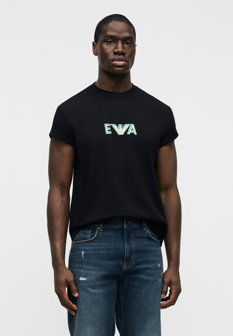 Μαύρο κοντομάνικο μπλουζάκι από βαμβάκι, με πράσινο κεντημένο λογότυπο "EVA" στο στήθος. Συνδυασμένο με μπλε τζιν με φθορές.