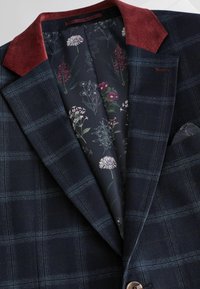 Marine checket blazer med maroon kordfløjskrave. Floral mønstret foring har mørk baggrund med farverige botaniske illustrationer.