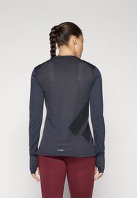 Dunkelblaues, langärmliges Sportshirt mit Mesh-Struktur, geometrischen schwarzen Akzenten und Daumenlöchern. Getragen mit weinroten Leggings.