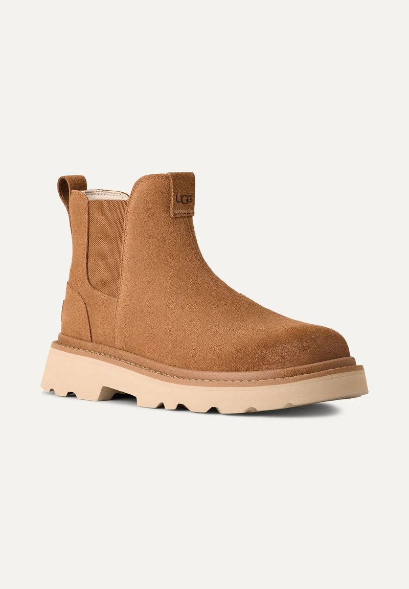 Brune ruskinds ankelstøvle med en flad beige sål, elastiske sidepaneler og en pull tab. Har et synligt brandlogo nær toppen.