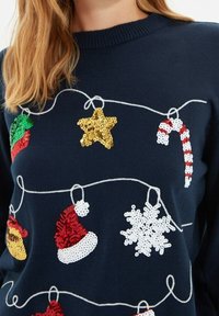 Navy Pullover aus Strick mit bunten Pailletten-Weihnachtsdekorationen: goldener Stern, rot-weißer Zuckerstange, Schneeflocke und Pompon-Akzenten.