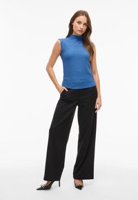 Blå texturerad ärmlös topp med hög hals, parat med svarta vida byxor och svarta spetsiga klackar med guld detaljer.