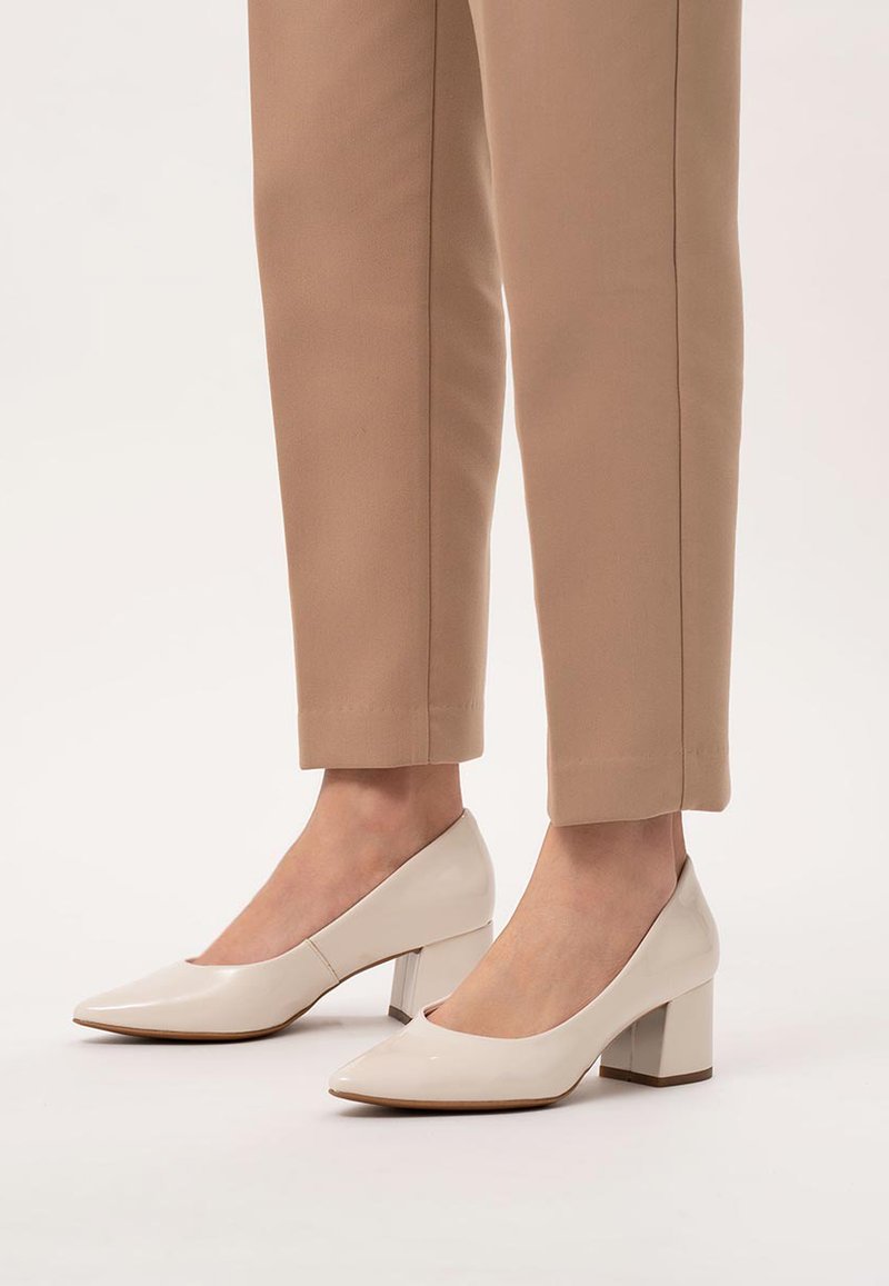 Beżowe lakierowane buty na słupku z ostrym noskiem, o gładkiej fakturze i stabilnym obcasie, w zestawieniu z beżowymi spodniami o wysokim stanie.