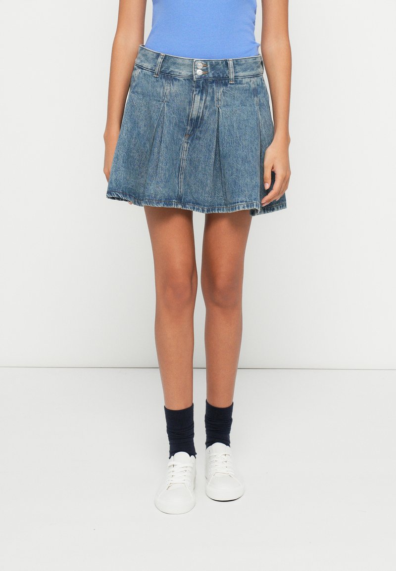 Fustă din denim cu design evazat, culoare albastru deschis, pliuri frontale și tiv sfâșiat. Asortată cu adidași albi și șosete scurte bleumarin.