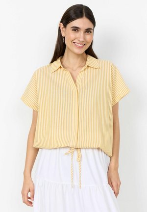 Uśmiechnięta kobieta z długimi ciemnymi włosami ma na sobie żółtą bluzkę w paski z krótkim rękawem, zawiązaną w talii, i białą, warstwową spódnicę.