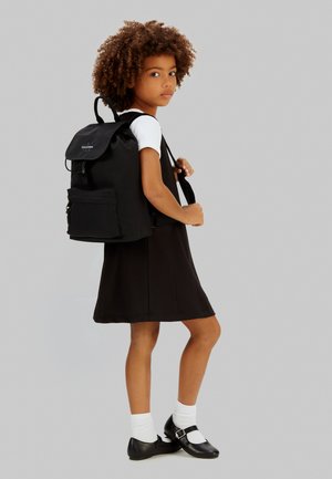 Ung pige med krøllet hår iført en sort kjole, hvide strømper, sorte sko og med en sort Calvin Klein rygsæk over den ene skulder.