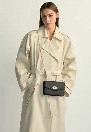 Młoda kobieta w beżowym trenczu, niosąca czarną skórzaną torebkę na ramię z złotym łańcuszkiem, na jednolitym tle.