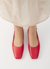 Ballerines en cuir rouge avec un design à bout carré. Texture lisse avec une finition brillante. Vue de dessus, partiellement couvertes par un vêtement de couleur claire.