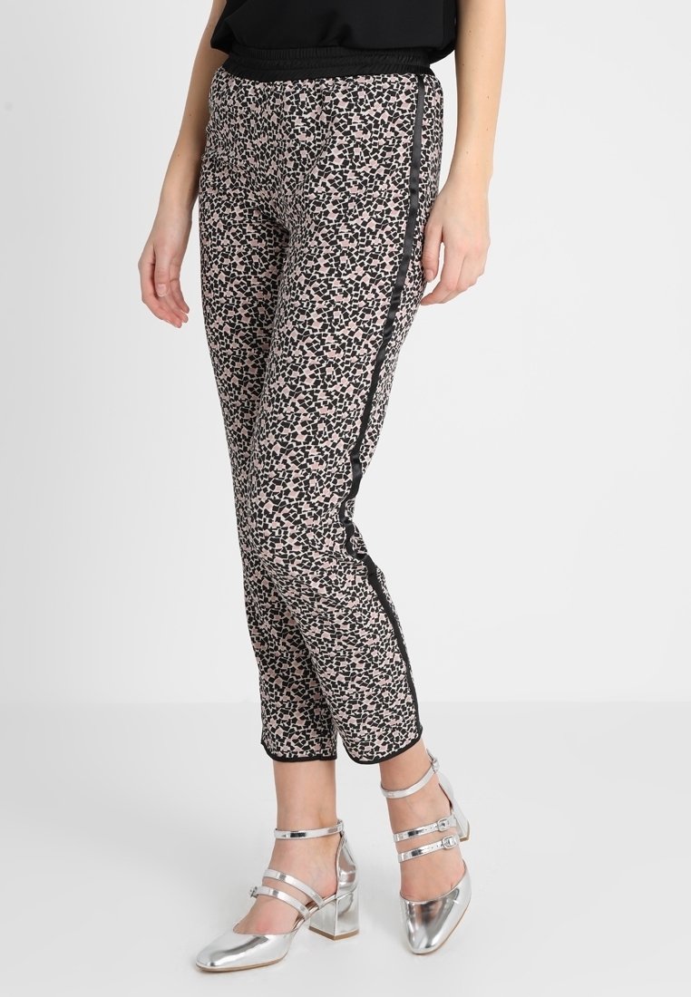 Femme portant un pantalon slim à motifs avec des bandes latérales noires et des chaussures à talons blocs argentés avec bride à la cheville, debout devant un fond uni.