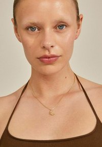 Collier en or avec un pendentif en forme de croissant de lune, porté sur une peau nue, associé à un haut sans manches marron. Textures lisses et design simple.