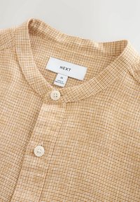 Chemise à manches longues à carreaux beige et marron avec col rond, deux boutons blancs et une étiquette de marque "NEXT", taille moyenne, fabriquée au Bangladesh.
