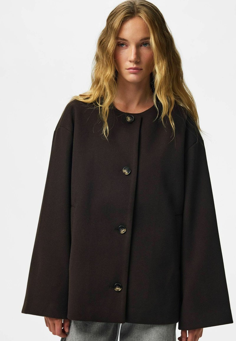 Femme aux cheveux blonds ondulés portant un manteau ample marron foncé boutonné et un pantalon gris sur un fond clair uni.