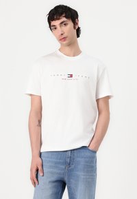 Bijela majica kratkih rukava s okruglim izrezom, s crvenim i plavim logo tekstom "TOMMY JEANS NEW YORK CITY" na prsima.
