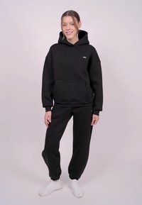 Sweat à capuche noir avec une poche frontale et un logo minimaliste, assorti à un pantalon de jogging noir et des baskets blanches.