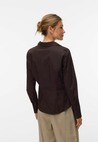 Chemise marron à manches longues avec un col classique et des poignets à boutons, associée à un pantalon beige clair, mettant en valeur un tissu lisse et structuré.
