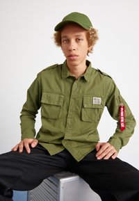 Green, long-sleeve shirt with two chest pockets and shoulder epaulettes, paired with black pants. A green cap and red zipper pull detail included.