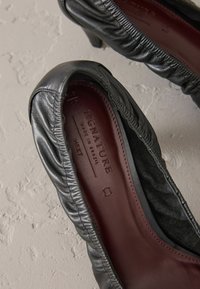 Schwarze Leder-Pumps mit einem gerafften Design, ausgestattet mit einer bordeauxfarbenen Innensohle, die "SIGNATURE" markiert ist, vor einem hellen strukturierten Hintergrund.