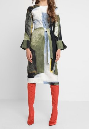 Vrouw draagt een jurk met lange mouwen met een abstracte print, wijde manchetten en een tailleband, gecombineerd met felrode kniehoge laarzen.