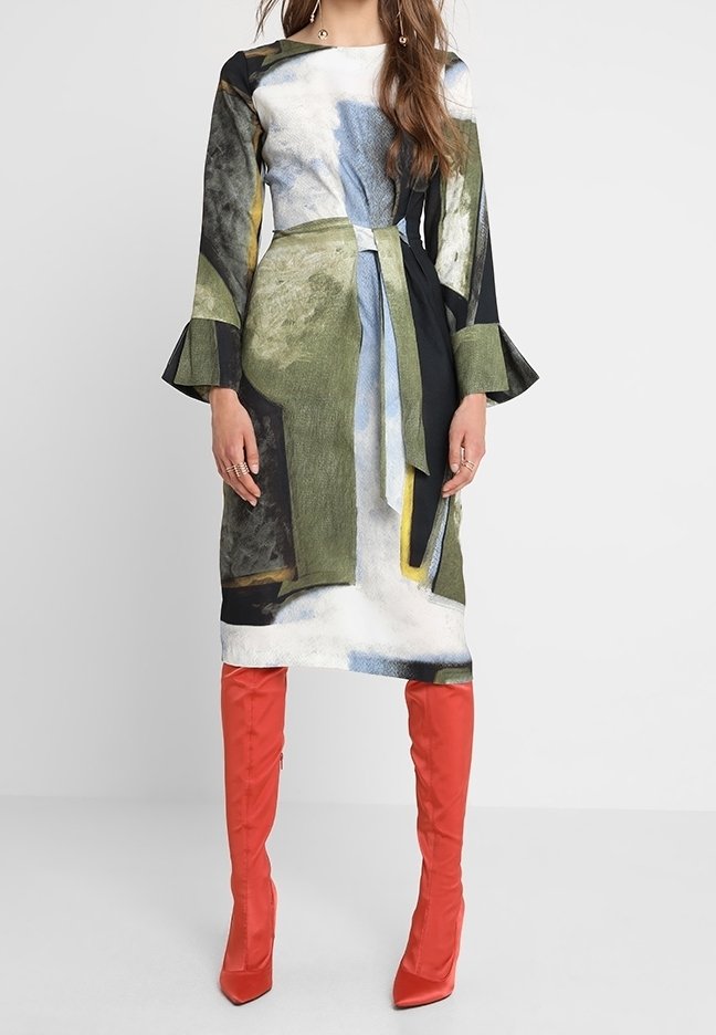 Femme portant une robe à manches longues à imprimé abstrait avec des poignets évasés et une ceinture à la taille, associée à des bottes hautes rouge vif.