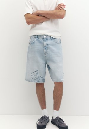 Personne portant un short en jean bleu clair avec une inscription noire délavée, des chaussettes blanches, des baskets gris foncé et une chemise blanche à manches courtes, bras croisés.
