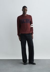 Burgunder strikkegenser med "GCDS" i mørkeblå, med hvite striper på ermene; kombinert med løse svarte bukser og svarte sko.