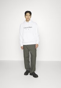 Witte hoodie met zwart "Calvin Klein" logo, gecombineerd met olijfgroene cargobroek en zwarte veterschoenen, staande tegen een witte achtergrond.