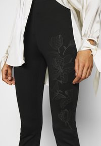 Zwarte leggings met een bloemenpatroon in een gedempte toon, gemaakt van rekbare stof, met een getailleerde pasvorm en gecombineerd met een witte blouse.