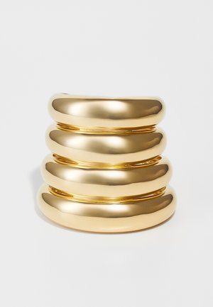 Anillo de metal color oro con un acabado suave y brillante. Diseñado con una forma curva apilada, presenta cuatro bandas redondeadas.