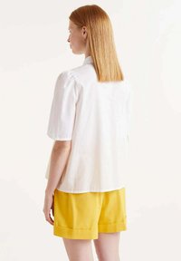 Camisa blanca de manga corta con un corte relajado, que presenta una textura suave. Combinada con pantalones cortos amarillos brillantes, con los puños doblados a la vista.