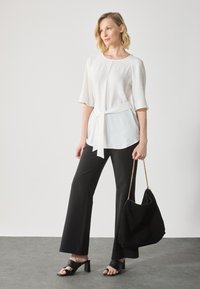Blouse blanche à manches courtes avec taille nouée, associée à un pantalon noir à pattes d'éléphant. Le mannequin tient un sac en daim noir et porte des sandales à talons ouverts noires.