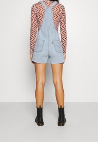 Pantalones de mezclilla azul claro con un ajuste relajado, que presentan tirantes traseros, dos bolsillos y dobladillo deshilachado, combinados con botas negras hasta el tobillo.