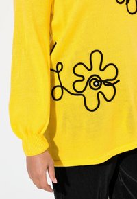 Gelber Strickpullover mit schwarzen Blumen- und Textmotiven, im lässigen Schnitt mit gerippten Bündchen, kombiniert mit schwarzen Cordhosen.