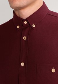 Man som bär en knäppt mörkröd skjorta med beige knappar och detalj vid kragen, vilket visar en del av bröstet och halsen.